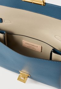 Blue leather handbag with beige interior, featuring a gold hardware clasp and a label stating "Coccinelle Authentic Italian Leather."