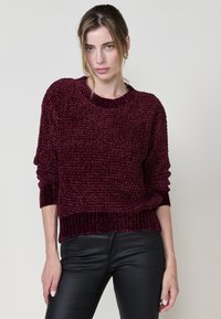 Burgunderfarbener strukturierter Strickpullover mit einem gerippten Rundhalsausschnitt und umgeschlagenen Ärmeln, getragen über schwarzen Lederhosen.