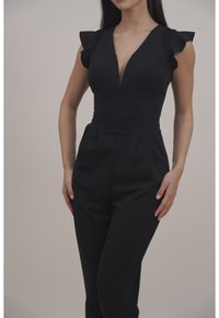 Svart jumpsuit med djupt V-ringat halsringning, korta volangärmar och spetsdetaljer på axlarna. Texturerat tyg och skräddarsydd passform.