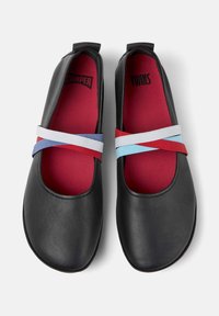 Zapatos de cuero negros con puntera redondeada, con interior rojo y correas elásticas cruzadas en blanco, azul y rojo en la parte superior.