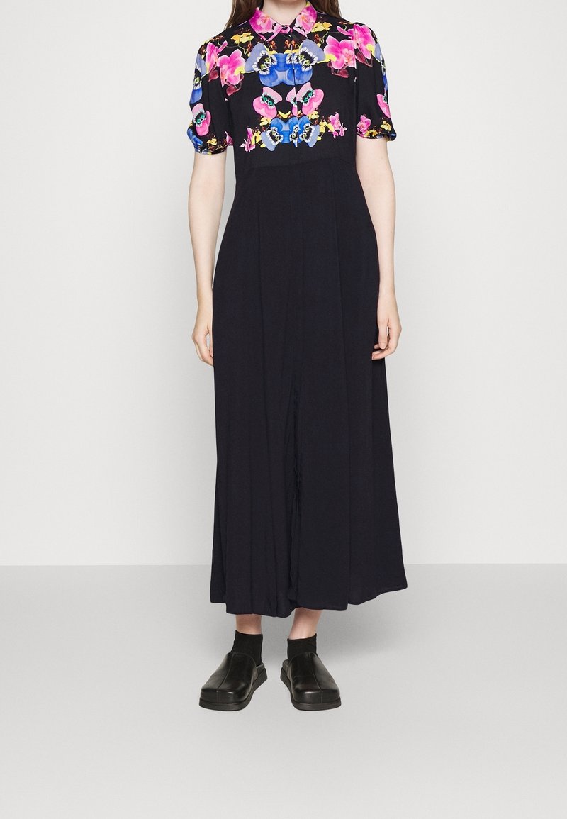Personne portant une robe midi noire avec des manches courtes et un col à motifs floraux roses et bleus, associée à des chaussures noires faciles à enfiler et des chaussettes.