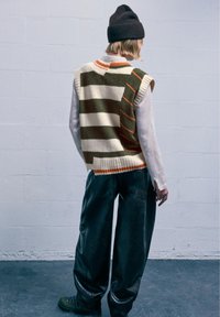 Gestrickte Weste mit grünen und cremefarbenen Streifen, orangefarbenen Akzenten. Kombiniert mit lockeren, grünen Kunstlederhosen und einer dunklen Mütze.