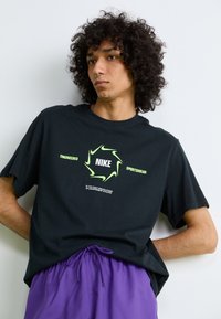 Pessoa jovem com cabelo encaracolado, vestindo uma t-shirt preta da Nike e calções roxos, de pé contra uma parede branca simples.