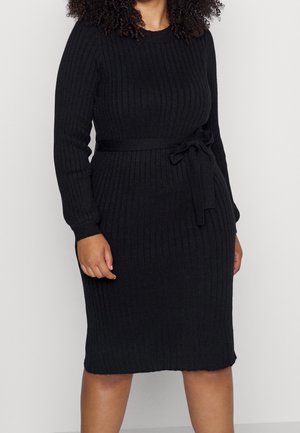 Kobieta w dopasowanej, długorękawowej, prążkowanej czarnej sukience dzianinowej z paskiem zawiązanym w talii, stojąca na tle jednolitego białego tła.