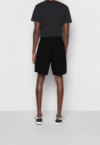 Person som bär svarta cargo-shorts, svart t-shirt och svart-vita Vans sneakers, står vänd bort mot en enkel bakgrund.