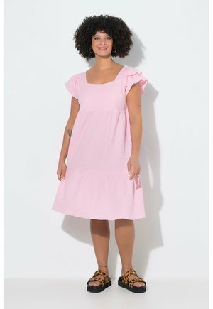 Nainen, jolla on kiharat hiukset, pukeutuneena vaaleanpunaiseen polvipituiseen mekkoon, jossa on liehuvat hihat ja leopardikuvioiset sandaalit, seisoo valkoista taustaa vasten.