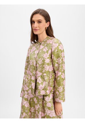 Femme portant une veste verte à motif floral rose et une jupe assortie, debout devant un fond blanc uni.