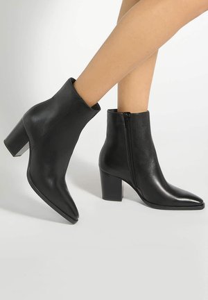 Bottines en cuir noir à talons blocs et fermetures éclairs sur les côtés, portées sur des jambes croisées sur fond blanc.