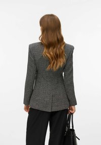 Szary żakiet o fakturowanej tkaninie z dopasowanym krojem, wyraźnymi ramionami i miękkim materiałem. Tył posiada centralne rozcięcie i jest zestawiony z czarnymi spodniami.