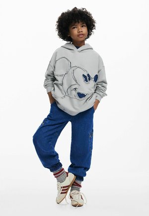 Hall halli kapuutsiga dressipluus, millel on suur kulunud efektiga Mickey Mouse'i kujutis, kombineeritud siniste cargo-pükstega. Jalatsid on valged punaste triipudega ning neid täiendavad triibulised sokid.