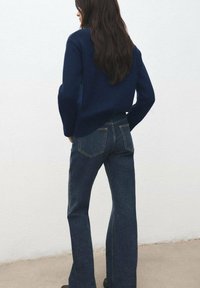 Temno modra pletena majica v kombinaciji s temnimi kavbojkami zvončastega kroja. Kavbojke imajo klasične zadnje žepke in tesno prilegajoč silhueto.