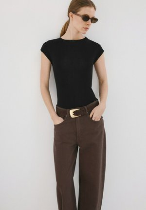 Vrouw in zwart shirt met korte mouwen, bruine broek met wijde pijpen en grote gouden gesp, draagt een zonnebril, staand tegen een witte muur met handen in de zakken.