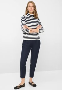 Gestreept top met lange mouwen in marineblauw en crème, gecombineerd met marineblauwe taps toelopende broek. Model draagt zwarte mesh ballerina's, armen gekruist, neutrale achtergrond.