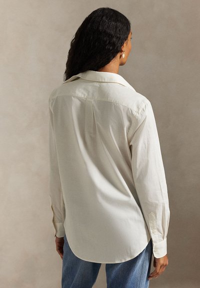 Femme aux cheveux bouclés foncés portant une chemise ample à manches longues écrue et un jean bleu, debout dos à un fond beige uni.