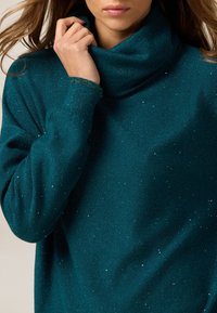 Türkisfarbener, glitzernder Rollkragensweater aus weichem Material mit lässiger Passform. Verfügt über einen dezenten Schimmer im gesamten Stoff und gerippte Bündchen.