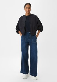 Schwarze Bomberjacke mit aufgeblähten Ärmeln, kombiniert mit hoch taillierten, weit geschnittenen blauen Jeans und weißen Sneakern. Einfaches, modernes Design.