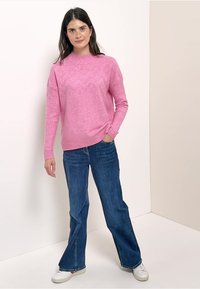 Jasnoróżowy sweter dzianinowy z teksturowanym wzorem, zestawiony z ciemnoniebieskimi dżinsami w stylu dzwonów i białymi trampkami. Modelka stoi na neutralnym tle.