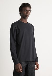 Ung mann med korte dreadlocks iført svart langermet skjorte og mørke jeans, stående mot en enkel lys bakgrunn.