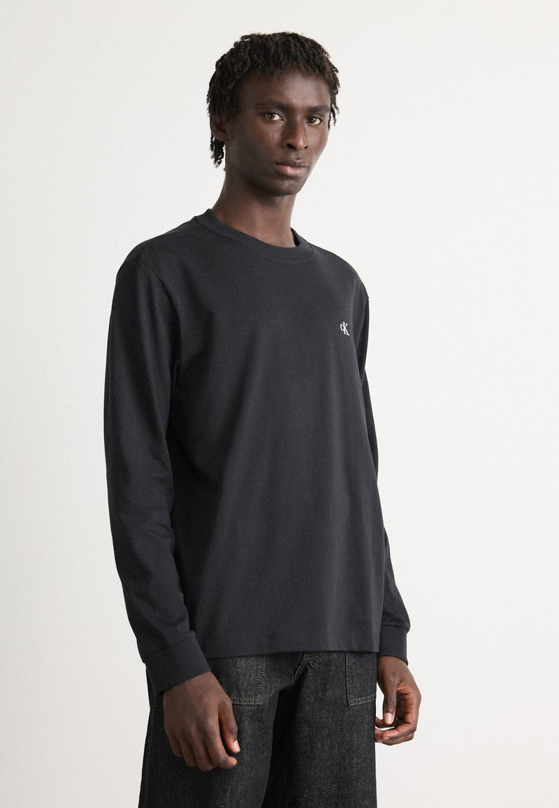 Giovane uomo con dreadlocks corti, vestito con una maglietta nera a maniche lunghe e jeans scuri, che si trova davanti a uno sfondo chiaro e semplice.