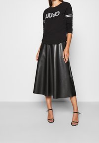 Donna che indossa un top nero a maniche lunghe con il logo "LIU JO", una gonna midi nera in ecopelle e sandali con tacco e cinturino alla caviglia neri.