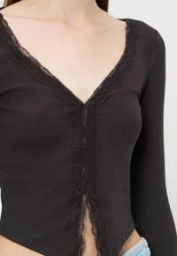 Gros plan d'un haut noir à manches longues avec une bordure en dentelle le long du profond décolleté en V et de l'ouverture frontale, porté avec un jean bleu clair.