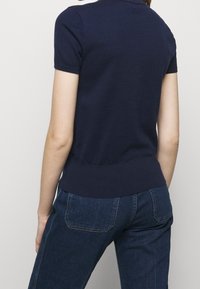 Mujer vestida con un suéter de manga corta azul marino y jeans oscuros, de pie de espaldas a la cámara contra un fondo claro y sencillo.