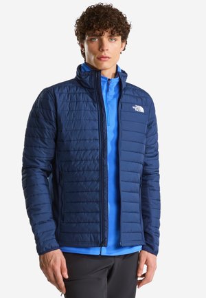 Mann mit lockigem Haar trägt eine marineblaue gesteppte Jacke über einem hellblauen Reißverschluss-Top und steht vor einem schlichten Hintergrund.