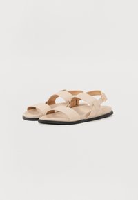 Sandales plates beiges avec des sangles entrecroisées, boucle réglable et texture lisse. Présente une semelle noire contrastante et un design minimaliste.