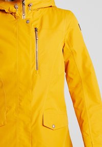 Veste imperméable jaune avec une poche poitrine zippée, une fermeture à boutons sur le devant et une capuche. Comprend des poches latérales et des accents métalliques.
