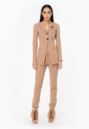 Traje beige a medida con una americana ajustada que cuenta con solapa de muesca y tres botones, combinado con pantalones ajustados y tacones con plumas.