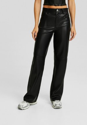 Persona con pantalones de cuero negros de tiro alto con un botón plateado, combinados con zapatillas blancas gruesas, de pie frente a un fondo gris.