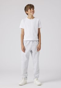 Witte katoenen t-shirt gedragen met lichtgrijze, elastische sweatpants; model draagt witte sneakers en staat op een neutrale achtergrond.