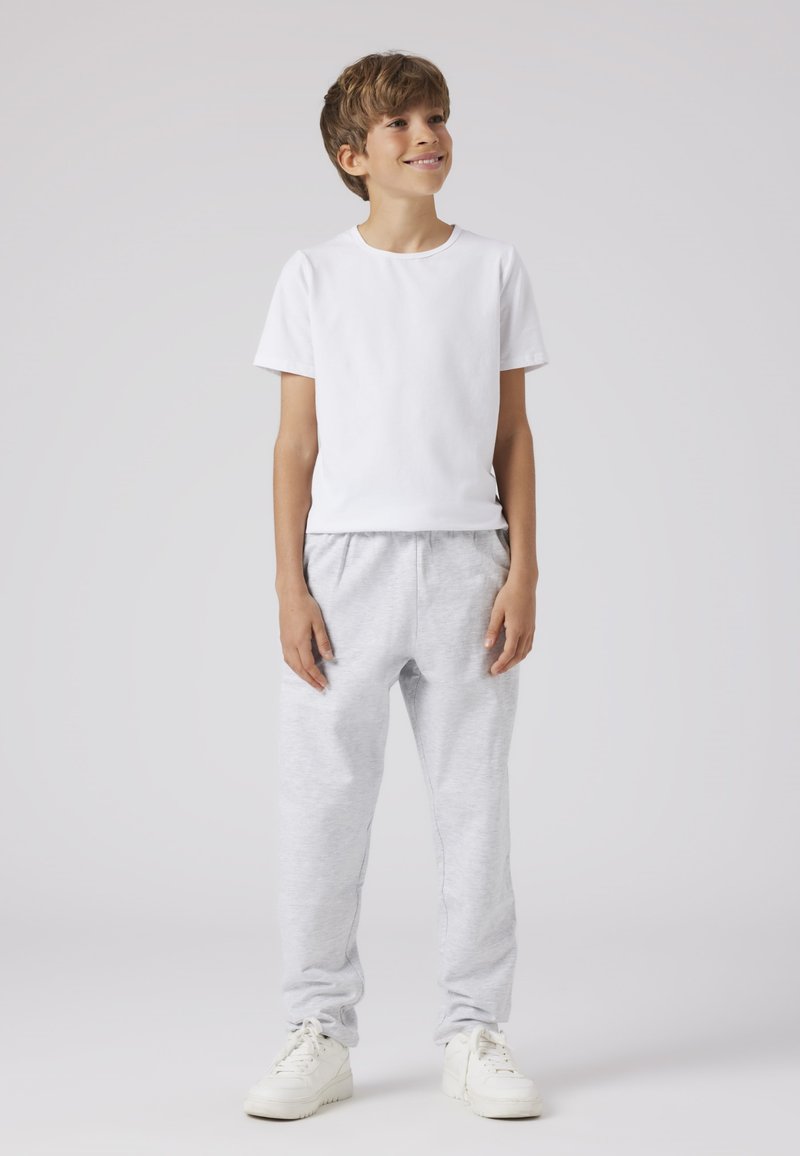 Witte katoenen t-shirt gedragen met lichtgrijze, elastische sweatpants; model draagt witte sneakers en staat op een neutrale achtergrond.