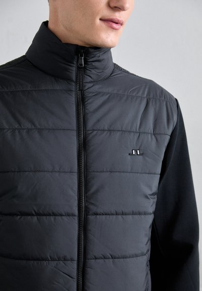 Black puffer vest with a high collar, horizontal stitching, and a front zipper. Features a small logo on the chest and a solid black fabric back.