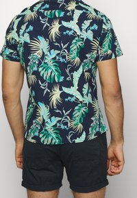 Kratký rukáv námořnické košile s barevným tropickým listovým vzorem, vyrobené z lehké látky, spárované s tmavými šortkami.