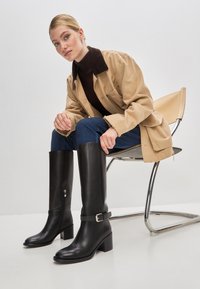 Cizme negre din piele până la genunchi, cu toc pătrat, având un fermoar lateral și un accent cu cataramă. Textură netedă și design elegant.