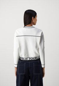Vit tröja med kontrasterande svarta ränder på ärmsluten och halsen. Har svart elastiskt midjeband med "KARLFELD"-text. Marinblå byxor med fickor.