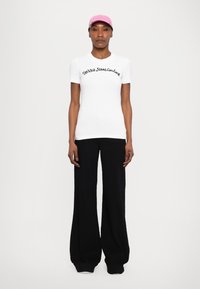White fitted t-shirt with black script text "Versace Jeans Couture," paired with wide-leg black pants and a light pink cap.
