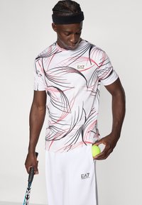 Λευκό αθλητικό πουκάμισο με κοντά μανίκια, διακοσμημένο με μαύρες και κόκκινες αφηρημένες γραμμές, με το λογότυπο EA7 στο στήθος, φορεμένο με λευκό παντελόνι, κρατώντας μια μπάλα του τένις.