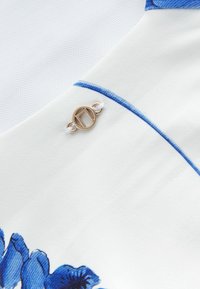 Biela látka s modrým kvetinovým vzorom, pričom obsahuje okrúhly zlatý gombík s logom. Hladký povrch a presné detaily stehu.