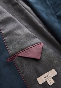 Innenfutter eines marineblauen Blazers mit gemustertem grauen Futter, bordeauxfarbenem Einstecktuch und Kleidungsetikett mit Marken-, Passform- und Größenangaben.