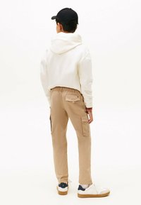 Witte hoodie, beige cargobroek met zijzakken en witte sneakers met donkere accenten. Model is van achteren gezien tegen een effen achtergrond.
