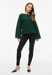 Maglione verde con texture e maniche ampie, abbinato a jeans skinny neri e décolleté nere a punta. Sfondo bianco pulito.