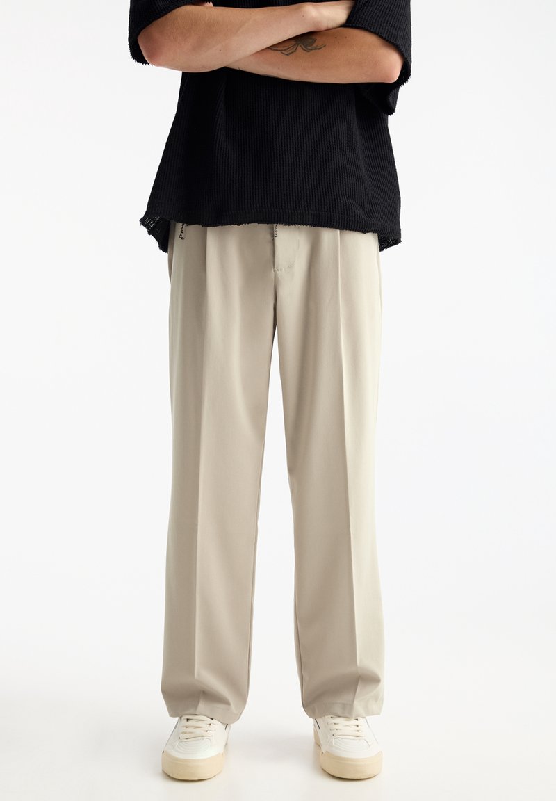 Personne portant un pantalon large beige, des baskets blanches et un pull noir, bras croisés sur un fond uni.