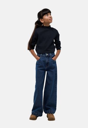 Un col roulé noir côtelé, un jean en denim bleu lâche à taille haute, porté avec des chaussures marron, les mains dans les poches, regardant vers le haut.
