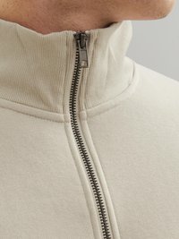 Beige Zip-Hoodie mit hohem Kragen, ausgestattet mit einem Metallreißverschluss und geripptem Stoff am Kragen, aus weicher Stoff gefertigt.