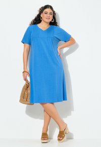 Niebieska sukienka z krótkim rękawem, dekoltem w kształcie litery V i detali z kieszeniami. W komplecie z plecioną torebką i brązowymi butami na platformie. Gładka faktura, casualowy design.