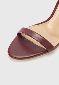 Chaussure à bout ouvert avec une sangle en cuir bordeaux et un intérieur nu. Présente une finition lisse et une forme de bout légèrement pointue.