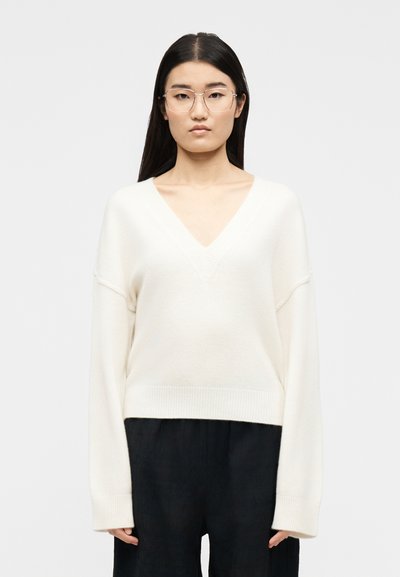 Jeune femme aux longs cheveux bruns et lunettes, portant un pull blanc à col en V et un pantalon noir, debout devant un fond clair uni.