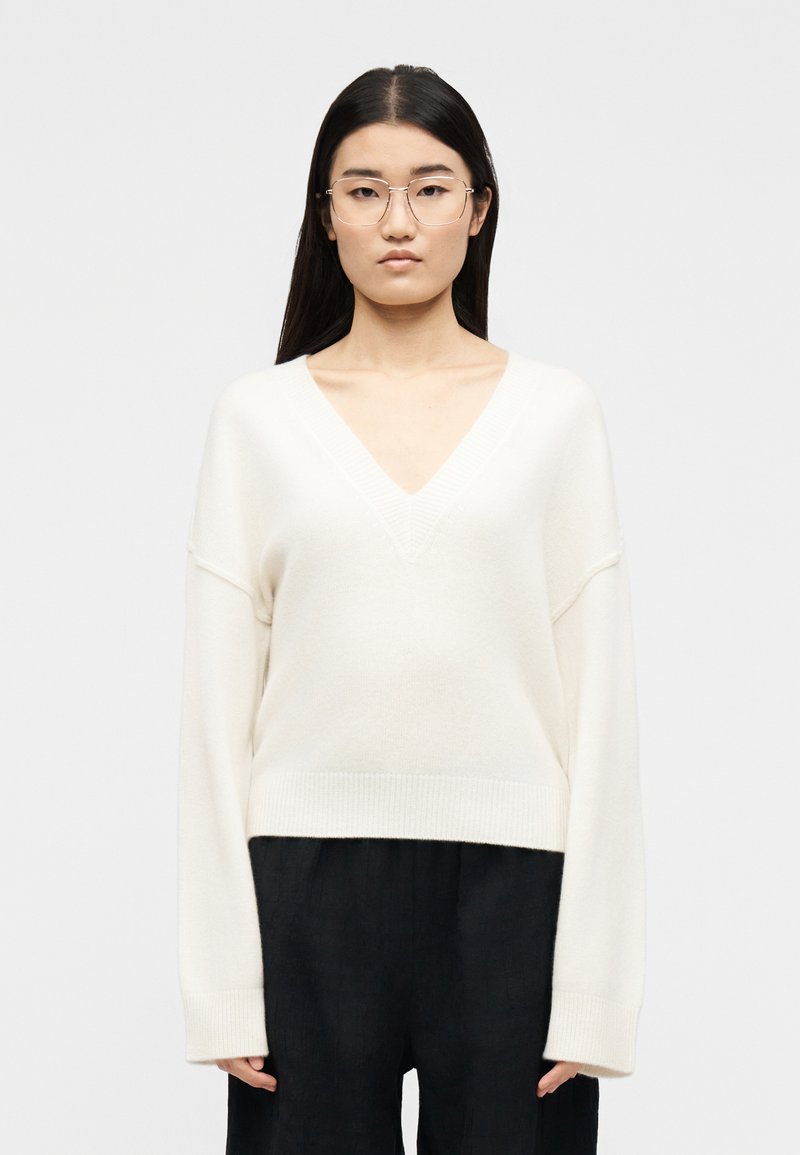 Νεαρή γυναίκα με μακριά σκούρα μαλλιά και γυαλιά, που φορά λευκό πουλόβερ με V λαιμόκοψη και μαύρο παντελόνι, στεκόμενη μπροστά σε απλό ανοιχτό φόντο.